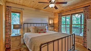 A wooden cabin bedroom with a bed, nightstands, a ceiling fan, and large windows with curtains.