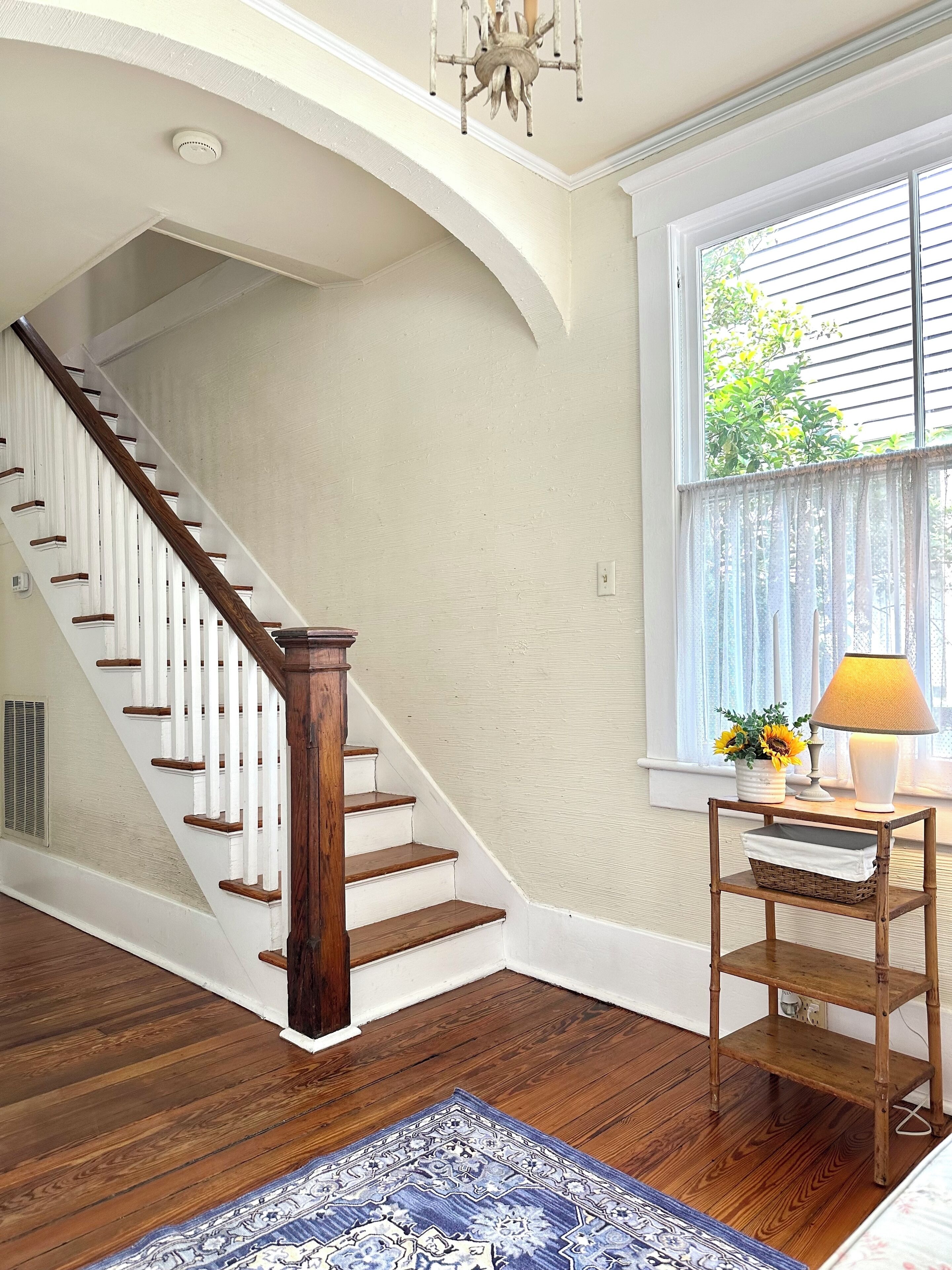 A staircase with wooden steps and white railings, a window with blinds, and a table with a lamp and flowers.