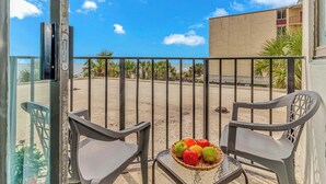 A balcony with two chairs, a table with a fruit basket, and a view of a building and palm trees. - Palace 105 (Myrtle Beach)
