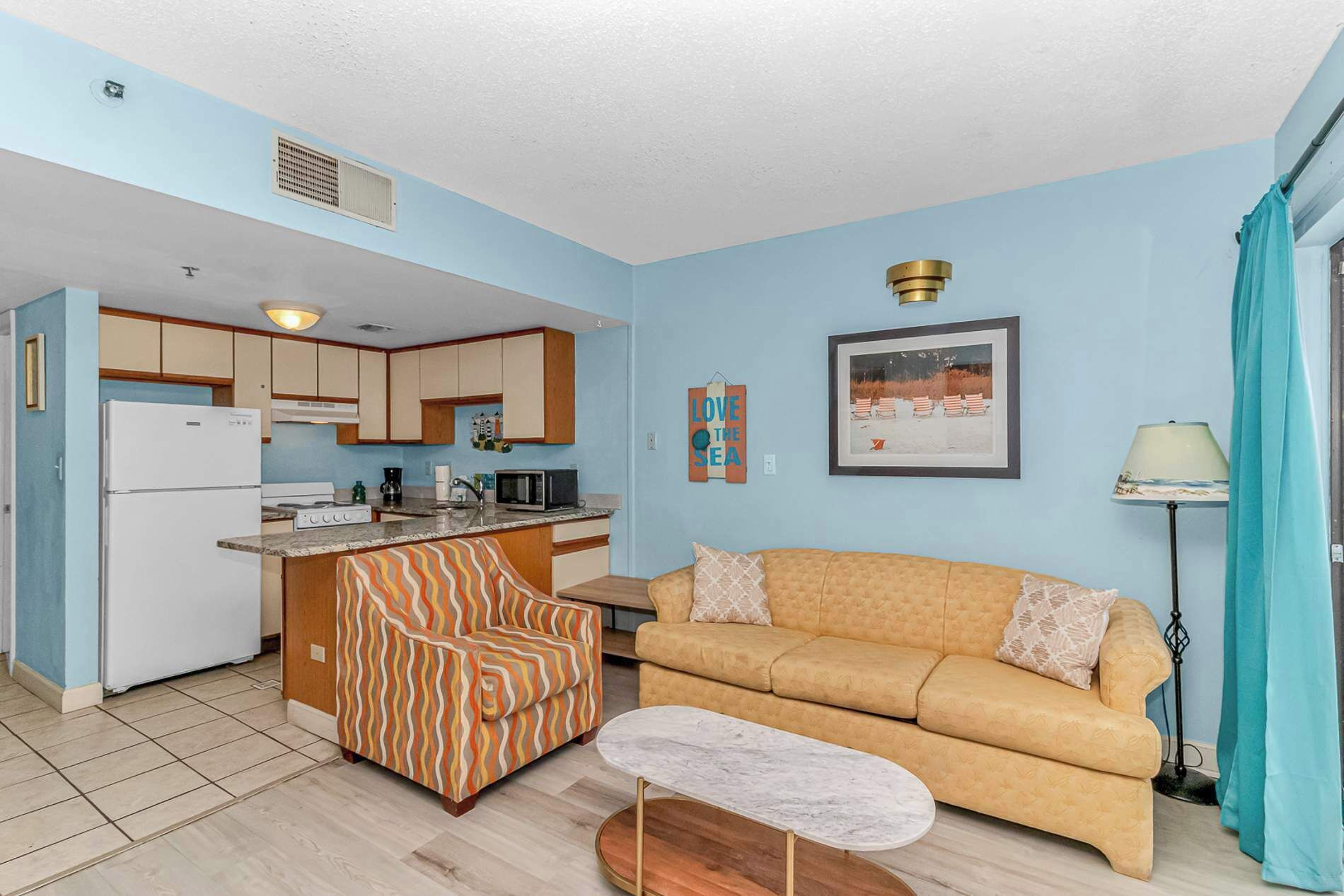 A compact living space with a kitchenette, a sofa, a patterned armchair, and a small table.