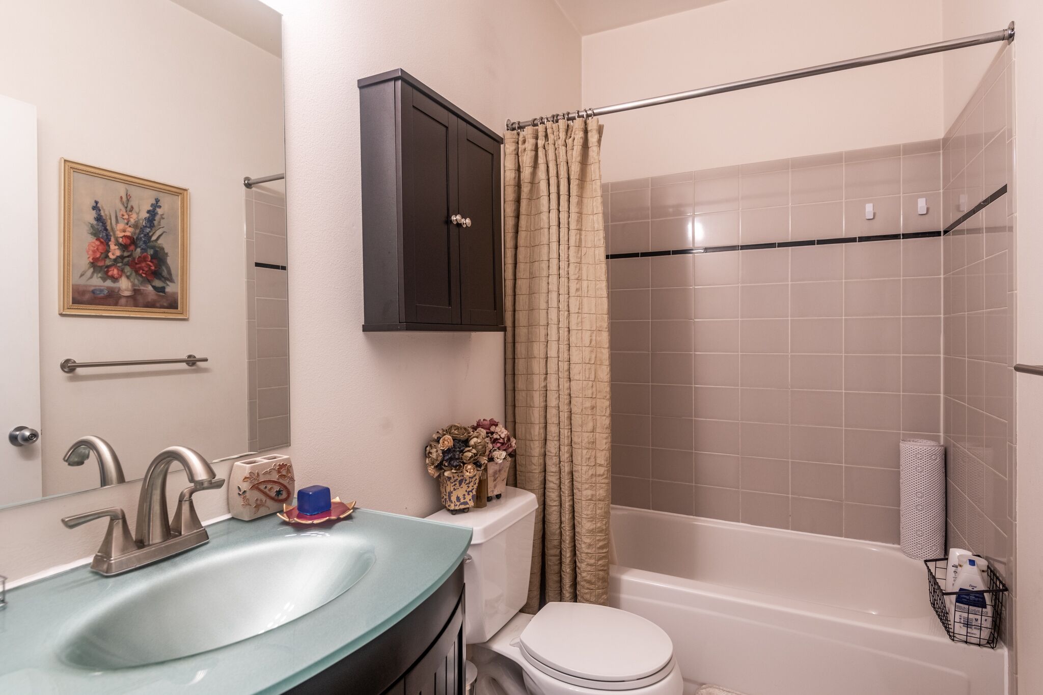 A bathroom with a glass sink, a toilet, a shower with a beige curtain, a dark cabinet, and a framed floral artwork on the wall.
