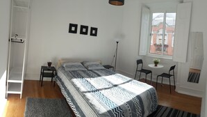 A bedroom with a bed, a wardrobe, a bedside table, a floor lamp, two chairs, a small table with a plant, and a window with a view of buildings. - Timor Guest House (Lisbon)