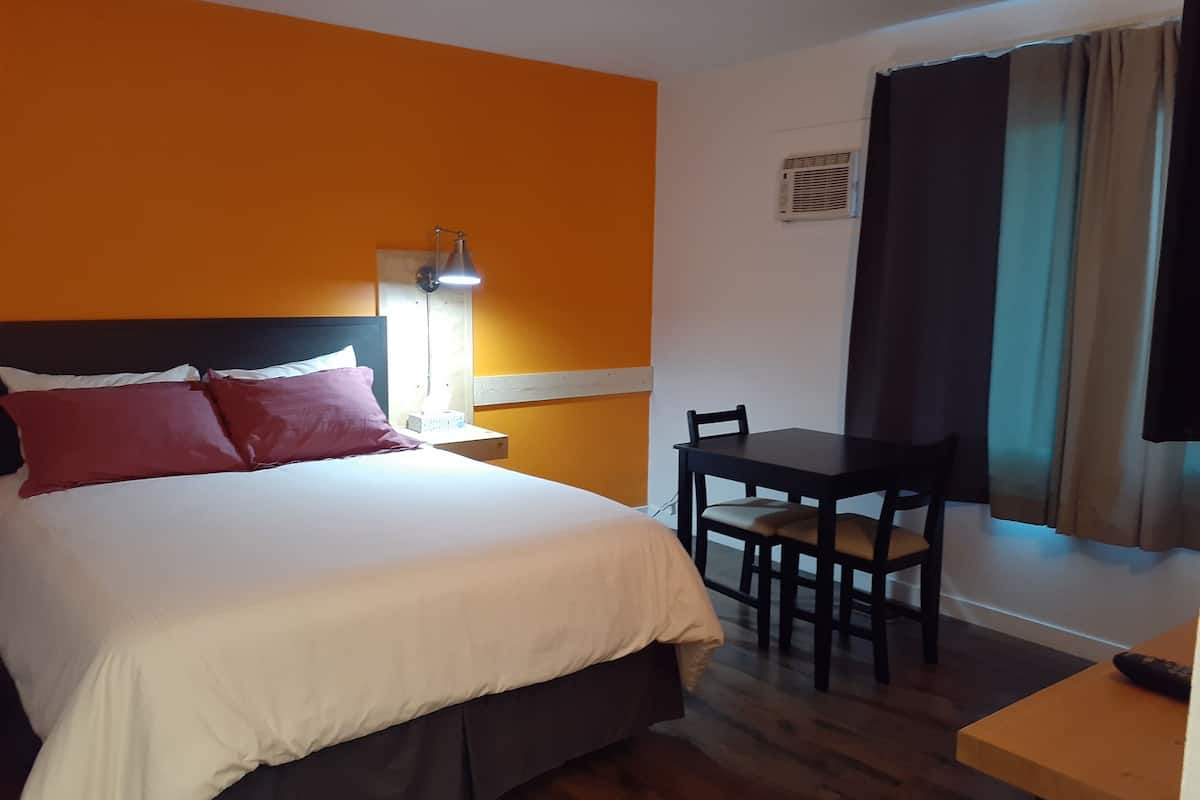 A hotel room with a bed, a desk, a chair, a nightstand, a lamp, and a window with curtains.
