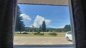 Un estacionamiento con un auto blanco estacionado al frente, un cielo azul despejado con nubes, y una vista de árboles y colinas al fondo.