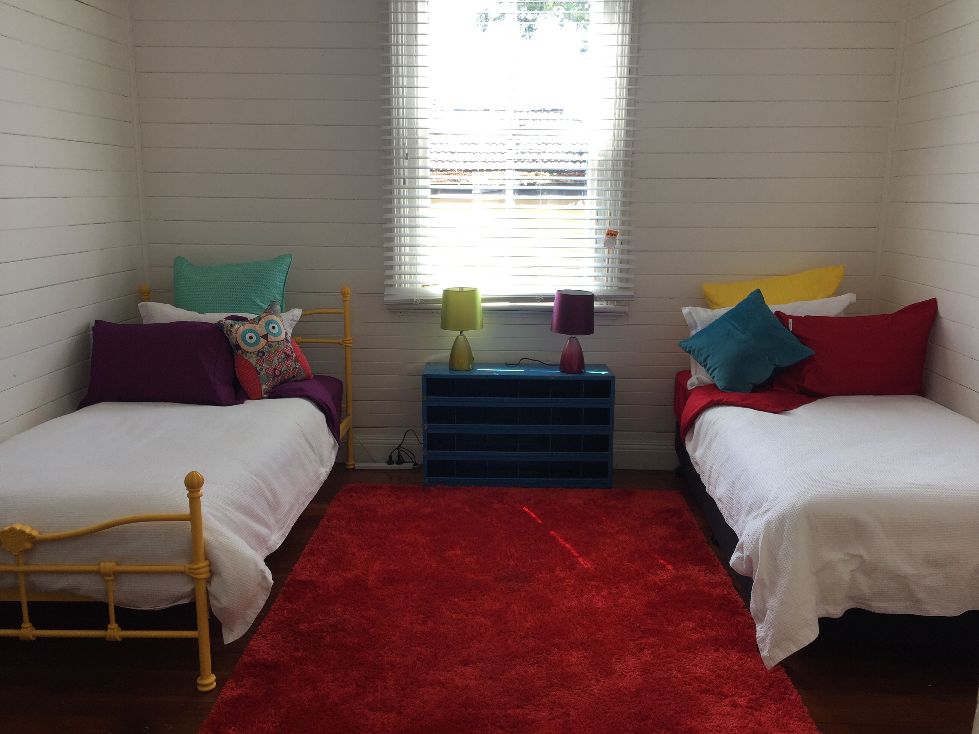 A room with two beds, a red rug, a blue nightstand with lamps, and a window with blinds.
