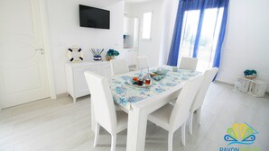 A dining area with a table set for four, white chairs, a TV mounted on the wall, and a window with blue curtains. - Pavon House Blue -Villetta 80 meters from the sea, 3 km from Torre San Giovanni beaches (Alliste)