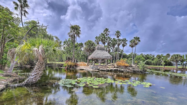 Um lago tranquilo com águas cristalinas, cercado por vegetação exuberante e uma construção com telhado de palha.