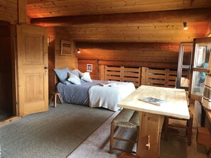 Um quarto acolhedor em estilo cabana, com teto de madeira, cama com roupa de cama azul, mesa de madeira com um banco e uma janela com cortinas.