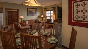A dining area with a wooden table set for four, surrounded by chairs. There is a sofa and a television in the background. - 2 Br: Soaring Eagle 211 (Snowshoe)