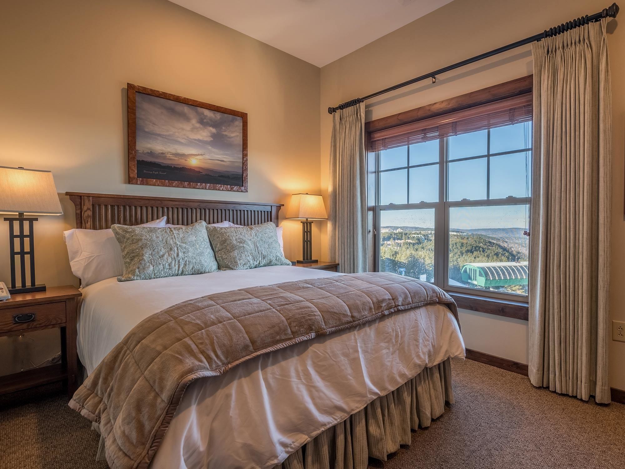 A hotel room with a large bed, two bedside lamps, a painting on the wall, and a window with curtains.