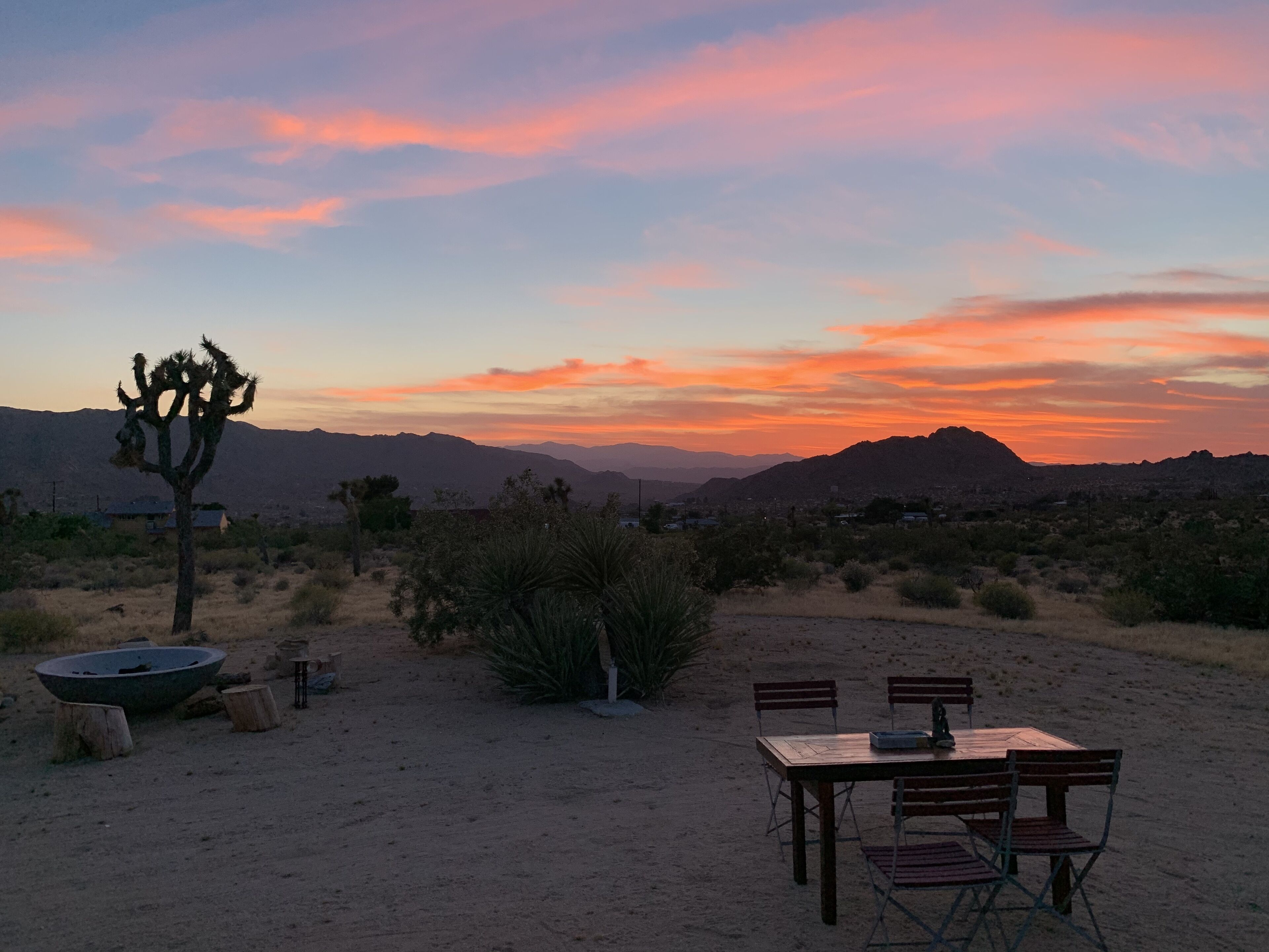 A desert landscape with a wooden table and chairs, a fire pit, and a cactus in the foreground.
