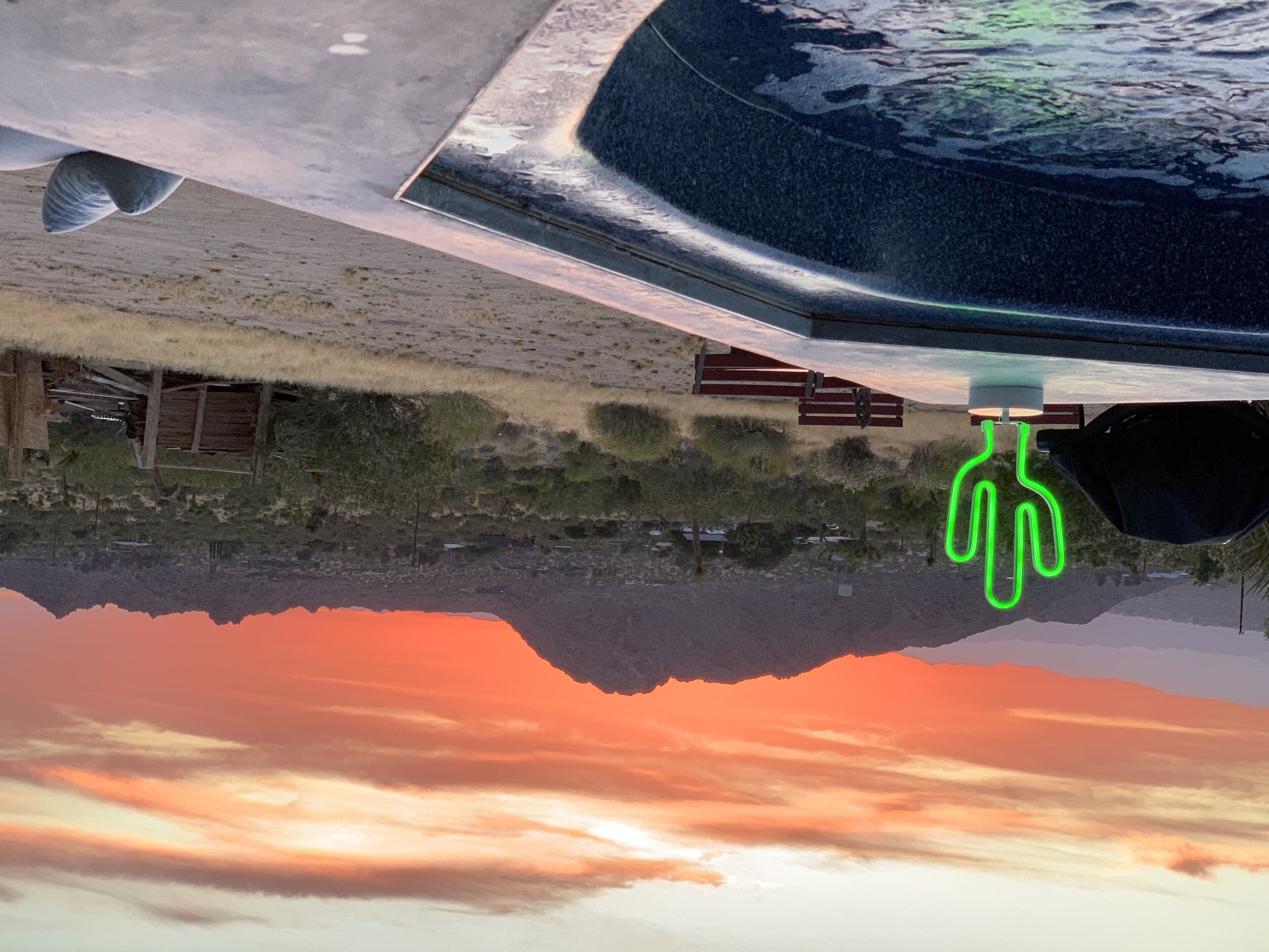 Le reflet d’un bâtiment avec un toit de chaume et des structures en bois, une enseigne au néon verte et un ciel au coucher du soleil.