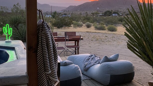 A desert landscape with mountains in the background, a wooden deck with a table and chairs, and a pool area with a neon cactus decoration. - Joshua Tree Monument Entrance Outside Your Front Door (Joshua Tree)