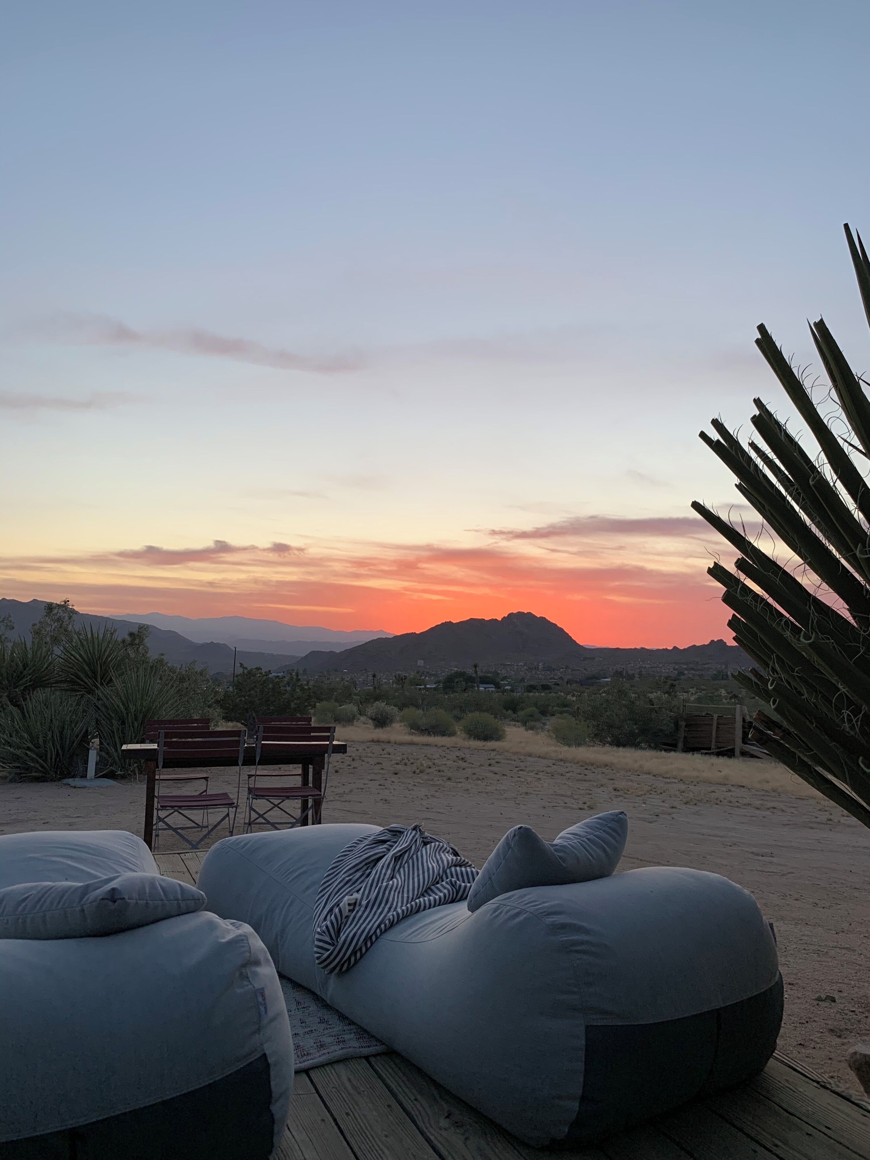 A wooden deck with large cushions and a striped blanket, overlooking a sunset over a desert landscape with sparse vegetation and distant mountains.