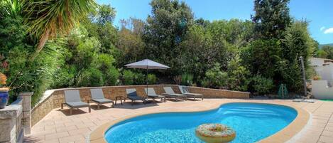 A swimming pool with lounge chairs and an umbrella, surrounded by lush greenery.