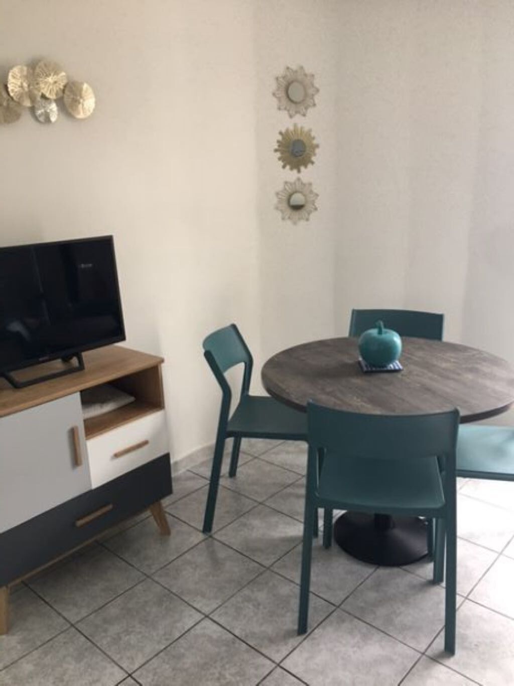 A dining area with a round table, teal chairs, a TV on a wooden cabinet, and decorative wall art.
