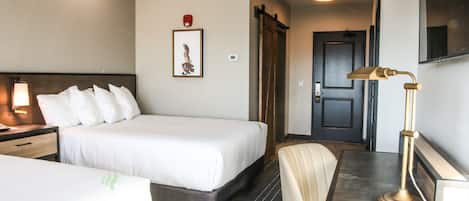 A hotel room with a bed, a desk with a lamp, a chair, a framed picture on the wall, and a door.