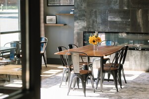 Uma sala de jantar moderna com mesa de madeira e cadeiras de metal, um vaso com flores amarelas e vista da rua através de grandes janelas.