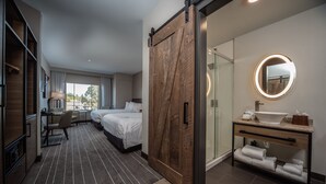 Habitació d'hotel amb una gran porta corredissa, un llit, un escriptori, una cadira, un bany amb un lavabo i un mirall, i vistes a l'exterior.