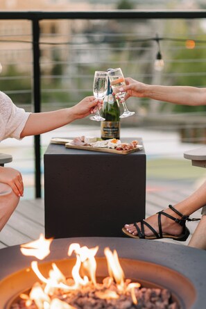 A fire pit with flames, a table with a bottle of wine and glasses, and a plate of food. - Hotel Windrow Downtown Ellensburg (Ellensburg)