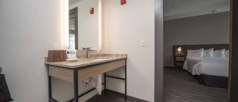 Ein modernes Hotelbad mit Waschbecken, Spiegel und Toilette. Der Raum verfügt über ein Bett mit weißer Bettwäsche und einen Nachttisch mit Lampe.