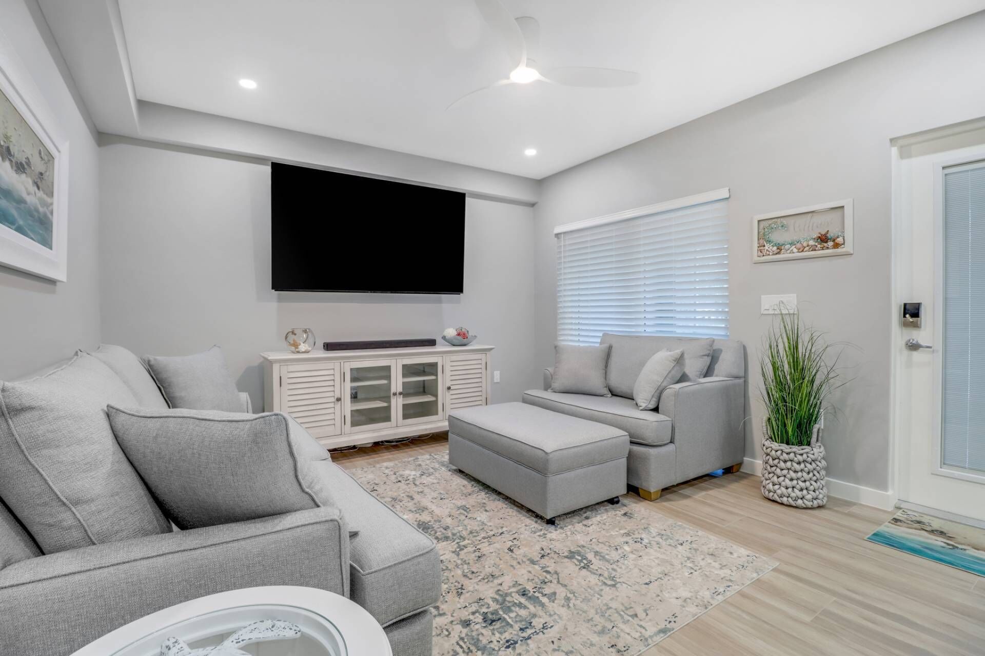 A modern living room with a grey sofa set, a flat-screen TV mounted on the wall, a white cabinet, and a large window with blinds.