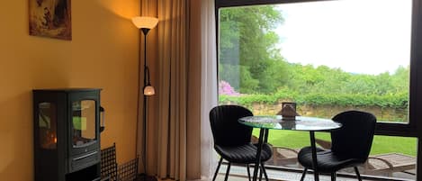 A room with a fireplace, a table with two chairs, a large window with a view of greenery, and a painting on the wall.