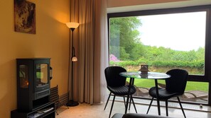 A room with a fireplace, a table with two chairs, a large window with a view of greenery, and a painting on the wall.