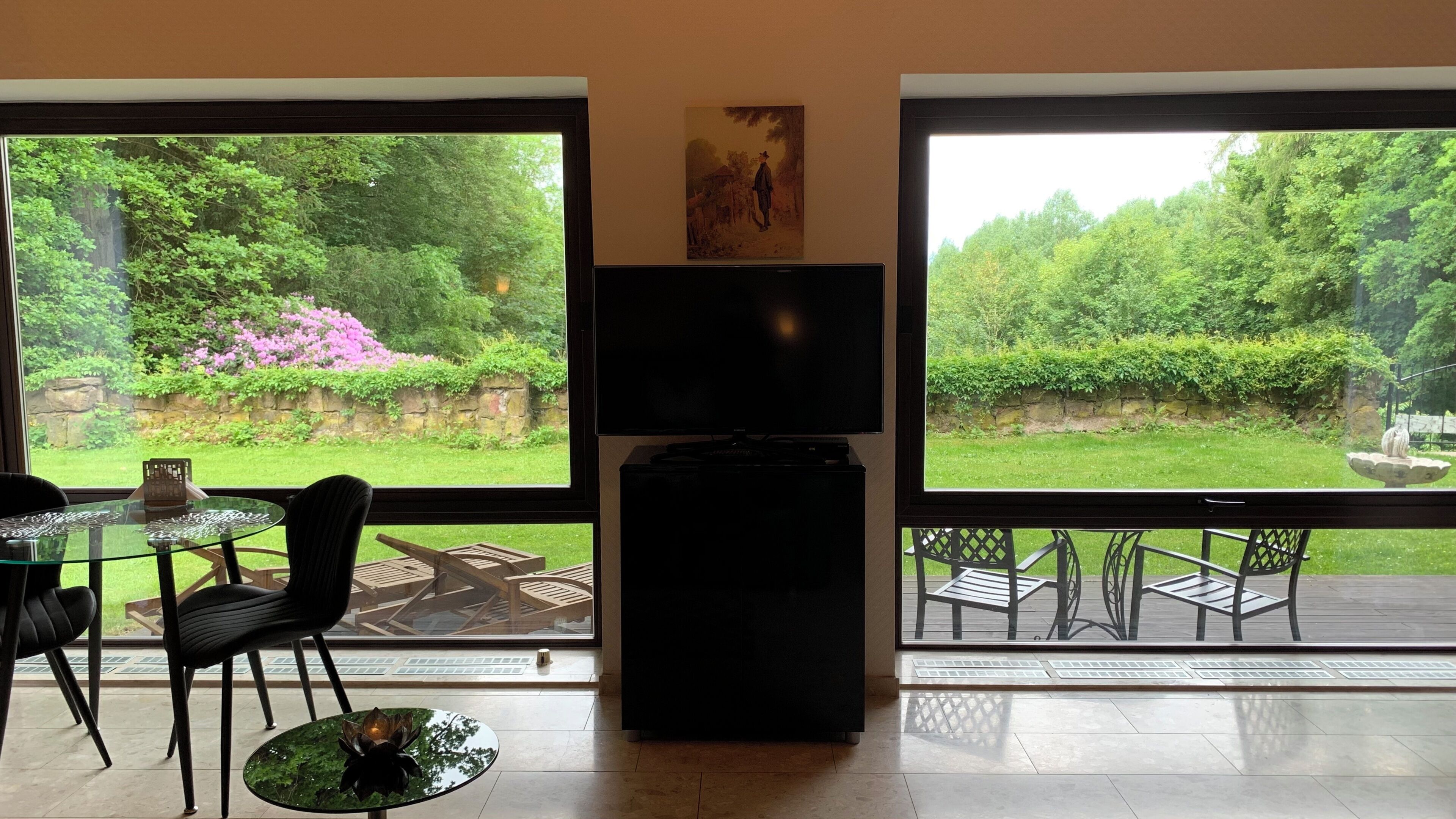 A room with a large window, a flat-screen TV, a dining table with chairs, and a statue in the garden view.