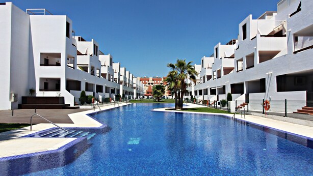 A swimming pool surrounded by modern white buildings with balconies, a clear sky, and a palm tree. - Al 317 - Penthouse near the beach with terrace (Vera playa)