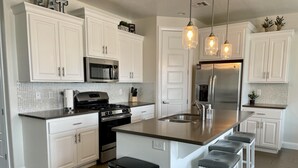 A modern kitchen with white cabinetry, stainless steel appliances, a central island with bar stools, and pendant lighting. - Sunny Ledgeview Retreat (St. George)