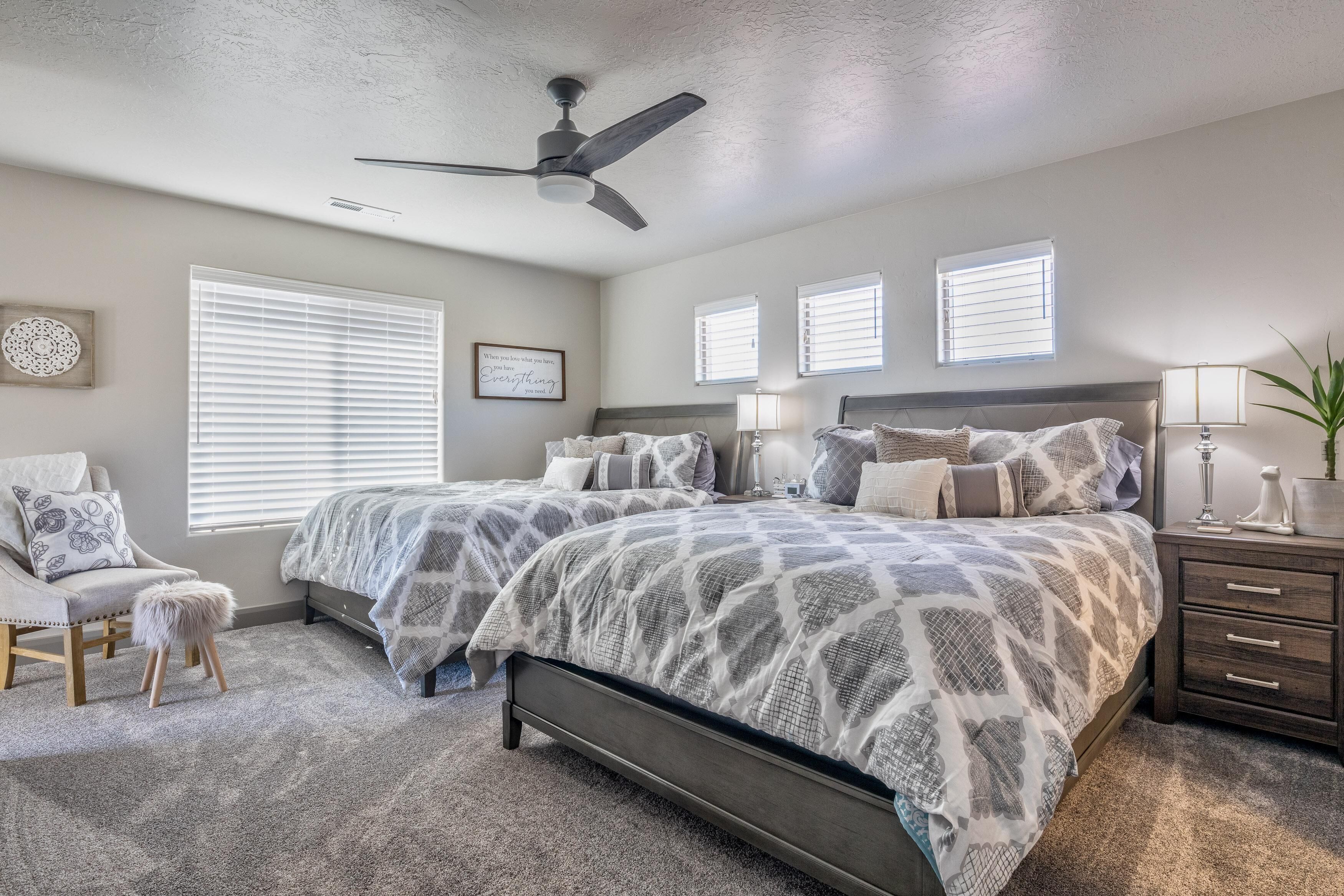 A bedroom with two beds, a ceiling fan, a chair, a bedside table, and a window with blinds.