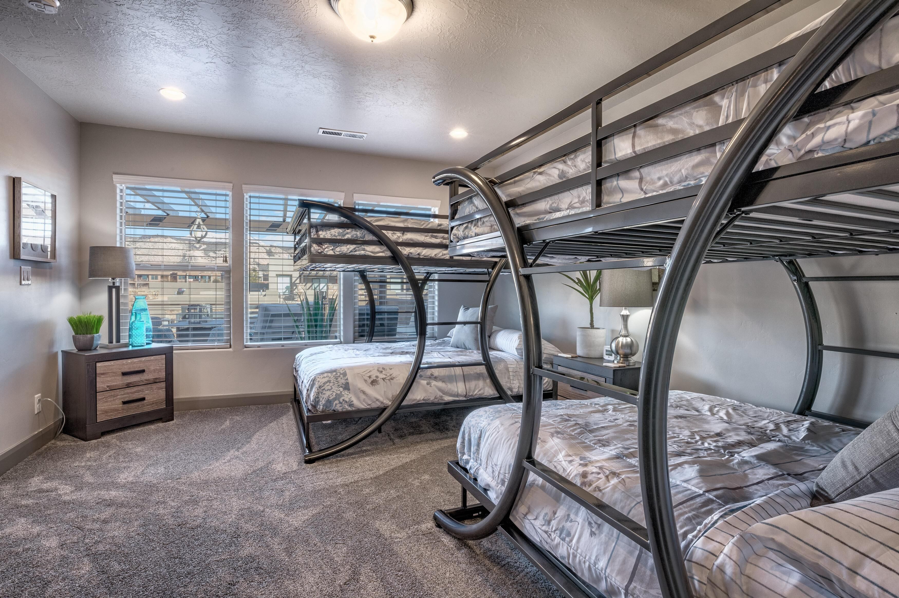 Dormitorio con literas y vistas a edificios a través de amplias ventanas, una mesita de noche con lámpara y una planta pequeña.