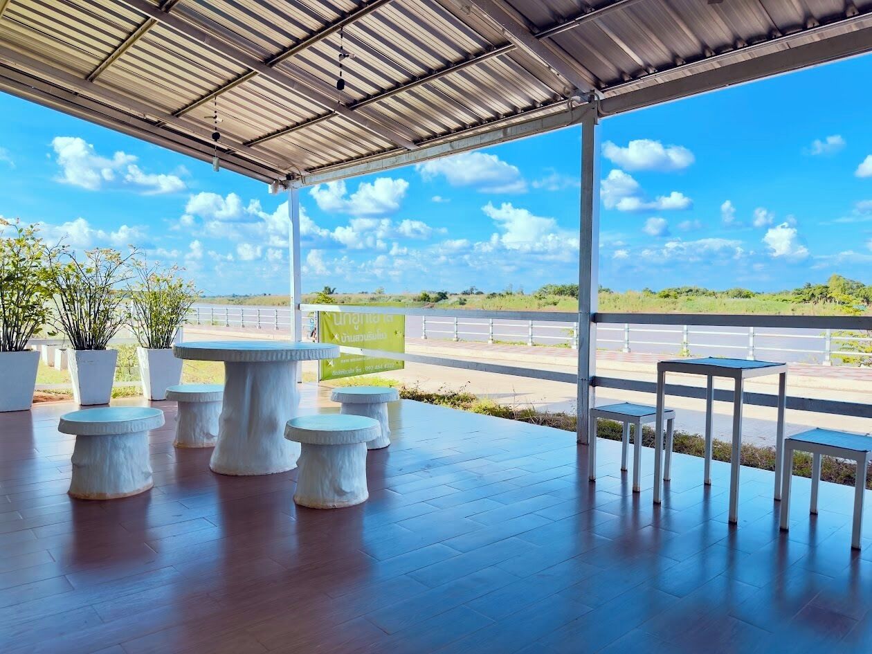 Une terrasse extérieure couverte, dotée de tables et de bancs en pierre, offrant une vue sur une route et sur la verdure, et surplombant un ciel bleu dégagé.