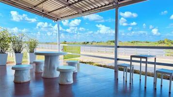 Une terrasse extérieure couverte, dotée de tables et de bancs en pierre, offrant une vue sur une route et sur la verdure, et surplombant un ciel bleu dégagé.