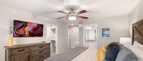 A bedroom with a bed, a dresser, a ceiling fan, a TV, and a bathroom visible through an open door.