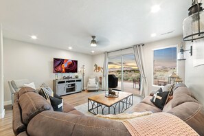 A modern living room with a large TV, leather couches, a wooden coffee table, and a view of the desert through large windows. - A Gathering Place (St. George)