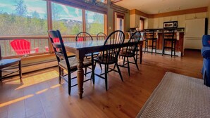Un comedor espacioso con mesa y sillas de madera, ventanas amplias con vistas a árboles y montañas, y una cocina con armarios blancos y un microondas.