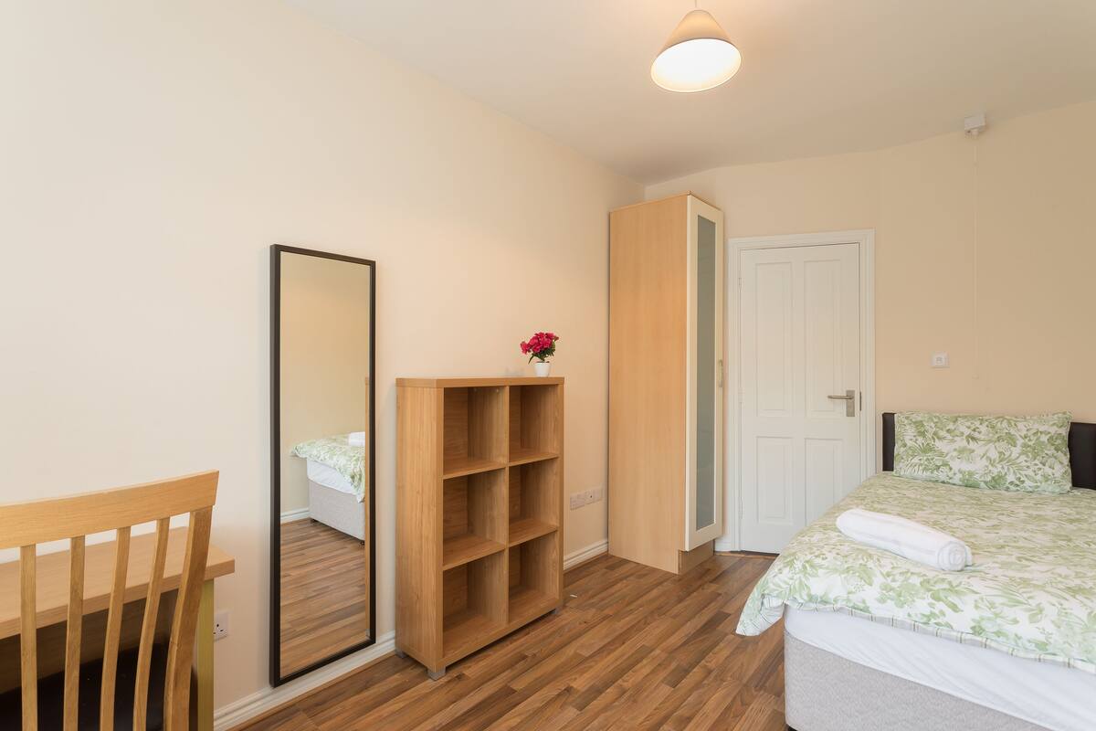 A bedroom with a wooden wardrobe, a mirror, a bed with a floral bedspread, and a wooden chair.