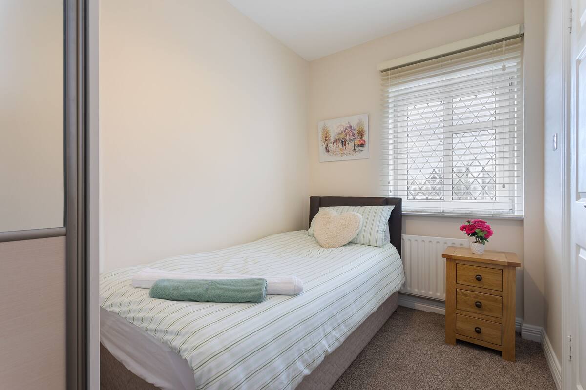A bedroom with a bed, a bedside table with a vase of flowers, a radiator, and a window with blinds.