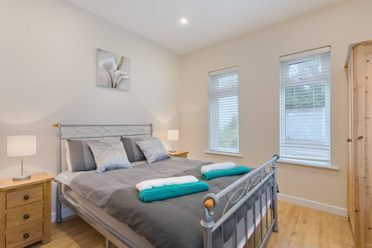 A neatly arranged bedroom with a metal-framed bed, two bedside tables with lamps, and two windows with blinds.