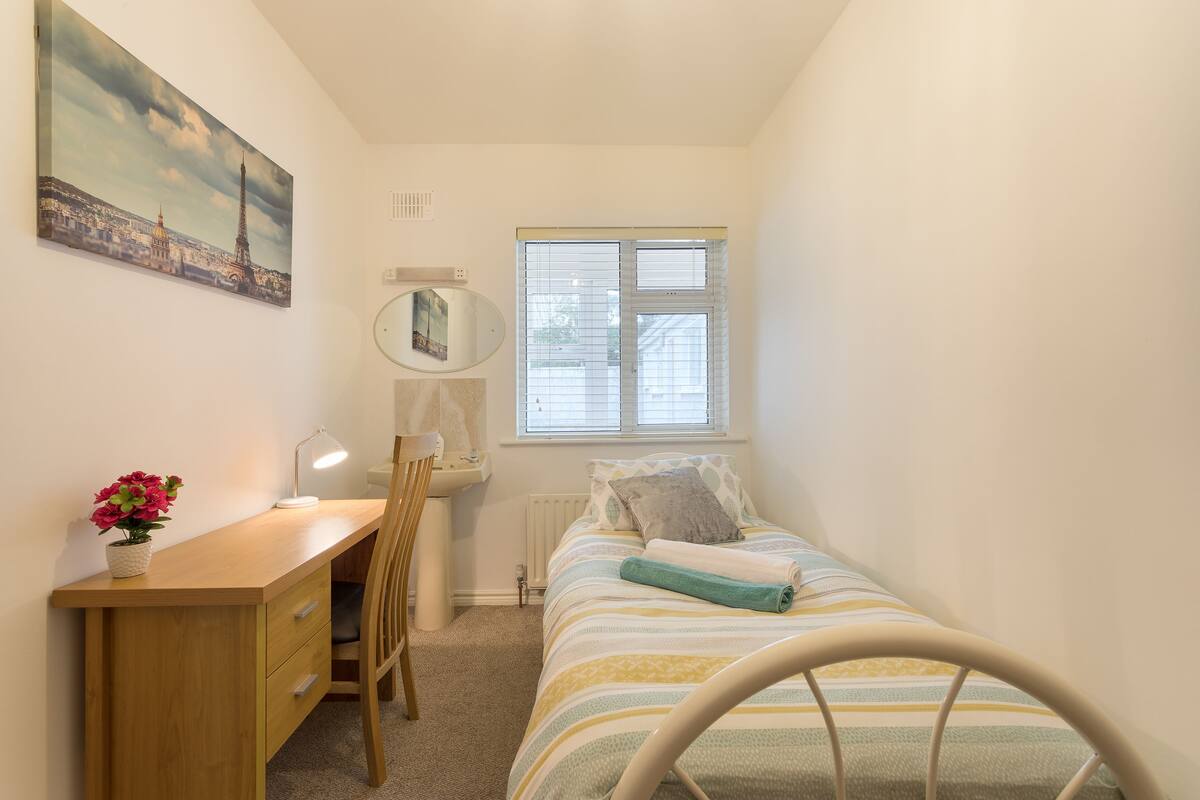 A bedroom with a bed, a desk, a chair, a radiator, a window, and a painting on the wall.