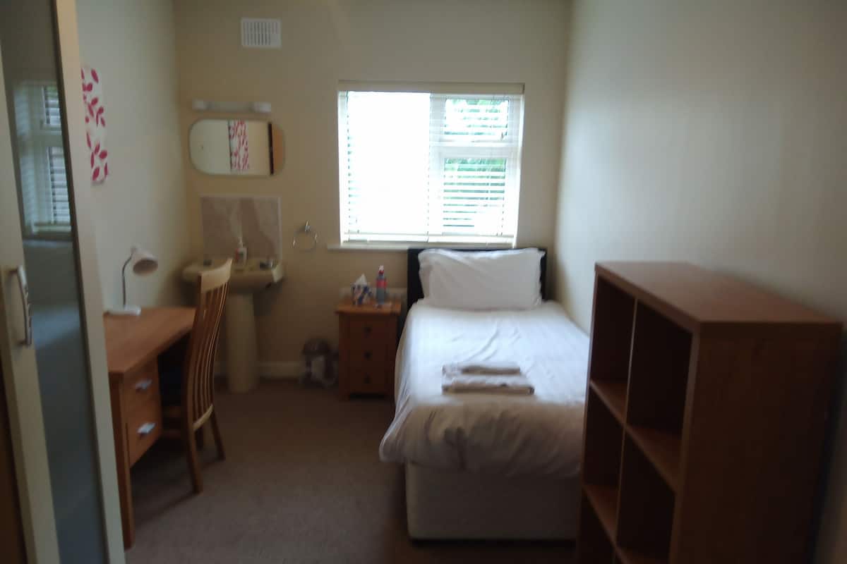 A bedroom with a bed, a desk, a chair, a bedside table, a window with blinds, and a radiator.