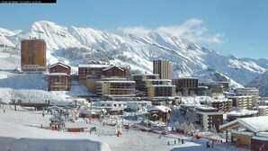 A ski resort with multiple buildings, including a tall hotel and several smaller lodges, set against a backdrop of snow-covered mountains.