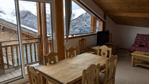 A wooden dining table with chairs, a television, and a sofa in a room with large windows overlooking snowy mountains.