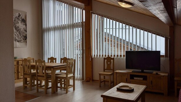 A dining area with wooden furniture, a television, and large windows with blinds.