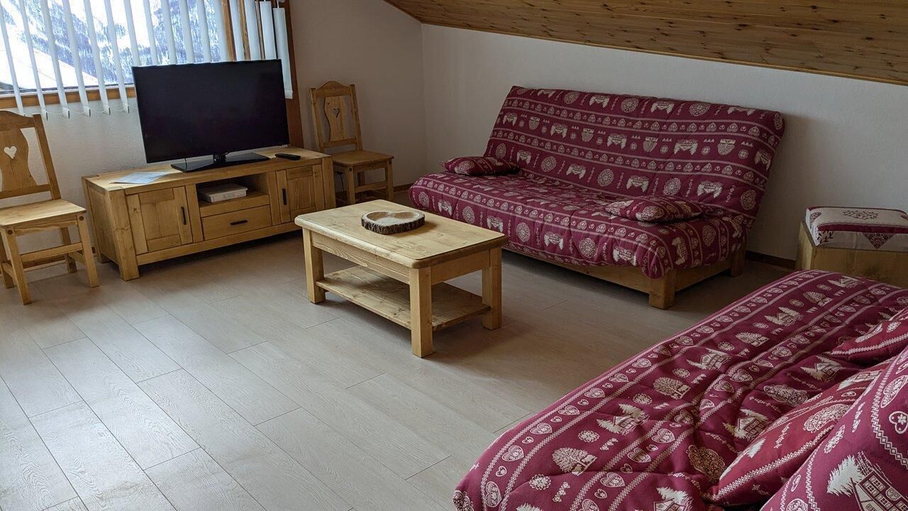 A cozy room with a wooden TV stand, a sofa with patterned cushions, a wooden coffee table, and a bed with a red blanket.