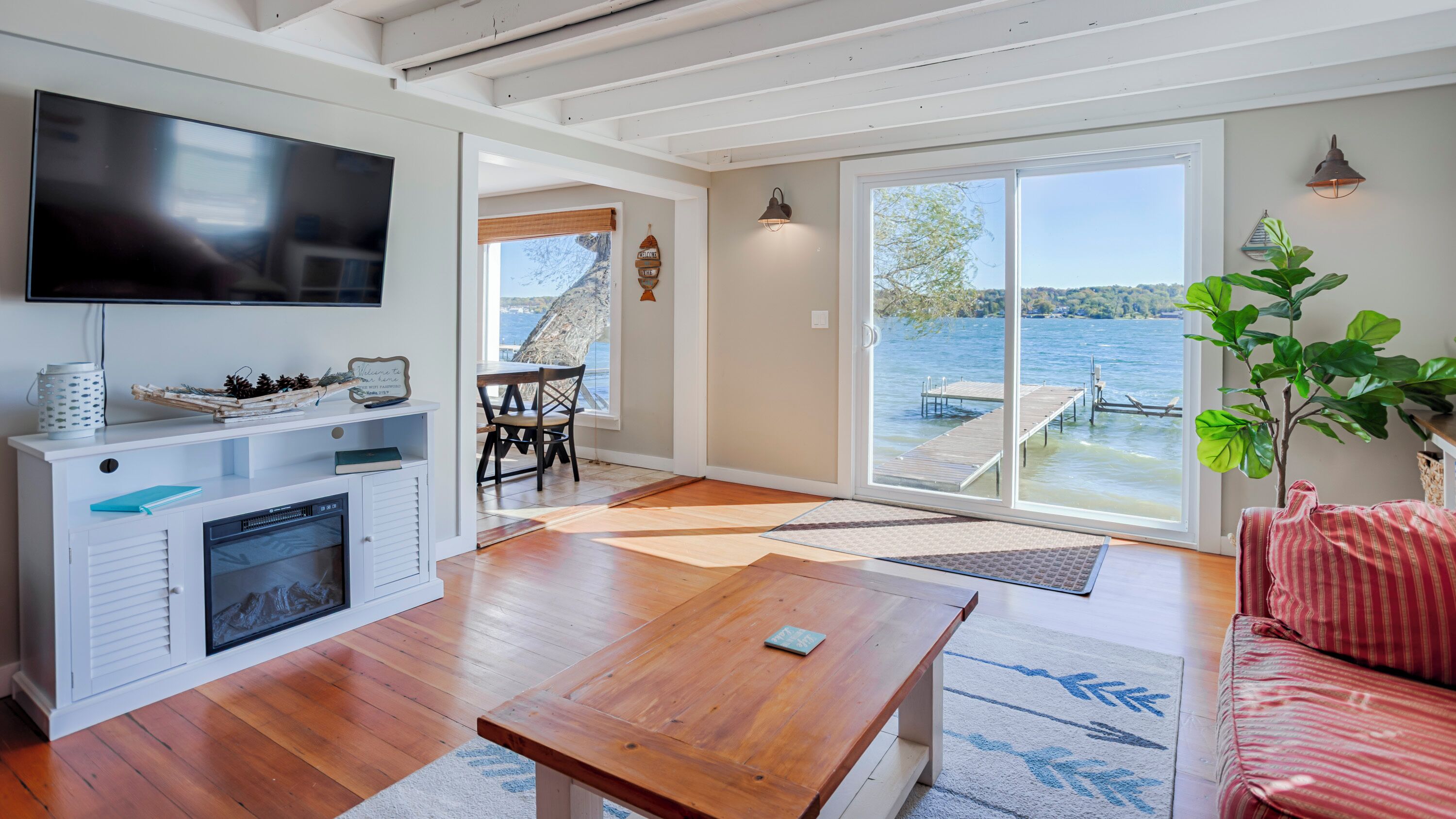 A spacious living room with a wooden floor, a flat-screen TV, a fireplace, a dining area with chairs, and a large sliding glass door leading to a dock by a lake.