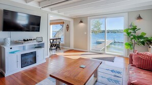 Amplia sala de estar con piso de madera, televisor de pantalla plana, chimenea, zona de comedor con sillas y un gran ventanal corredera que da a un muelle sobre un lago.