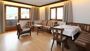 Ein gemütliches Zimmer mit Holzboden, einer Couch, einem Tisch und zwei Stühlen. Große Fenster mit Vorhängen.